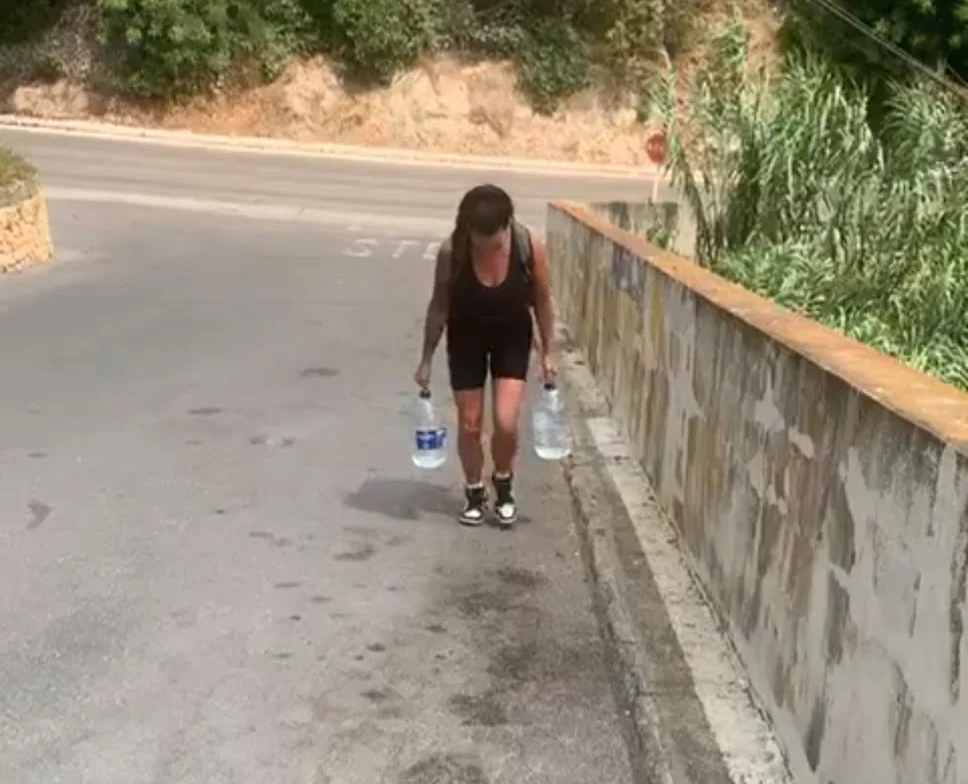 Woman in red sportswear carrying water bottles on mountain road.