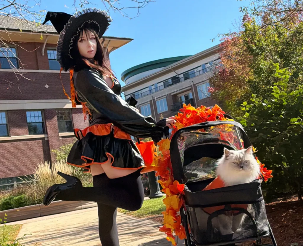 Woman in black costume posing with leg up beside Halloween-decorated cat pram
