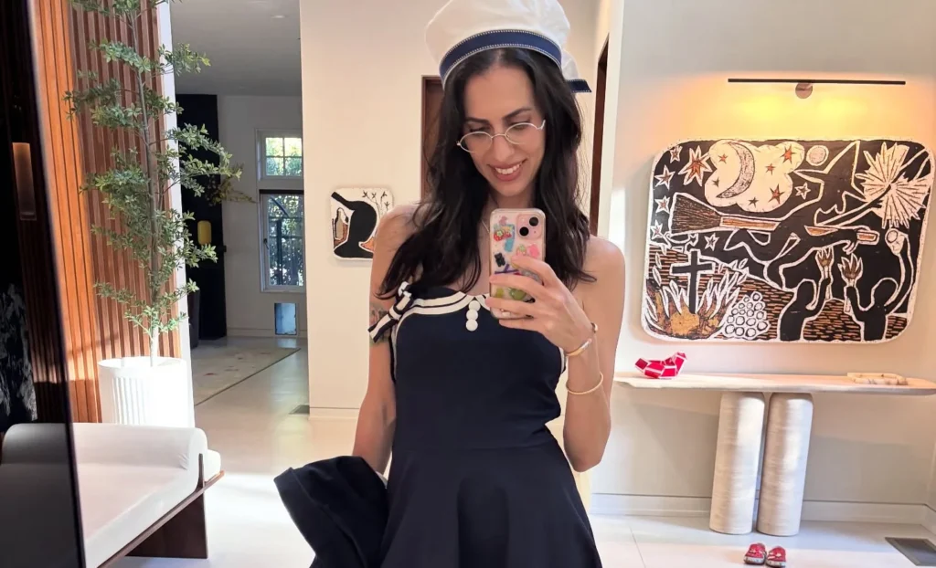 Hila Klein in black dress with white trim and white headband, posing indoors