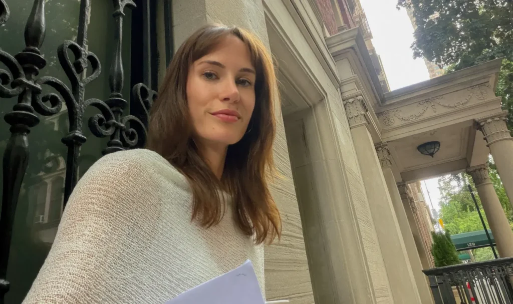 Woman in cream cardigan holding paper at ornate architectural entrance.