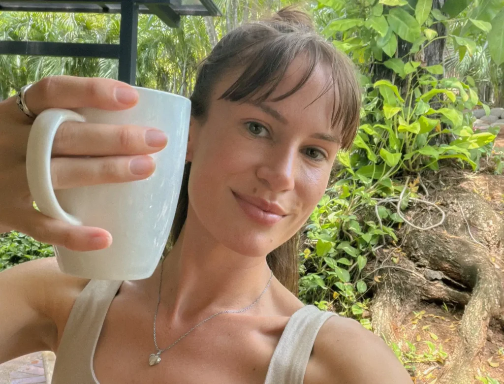 Woman in cream tank top holding white cup outdoors in garden setting.