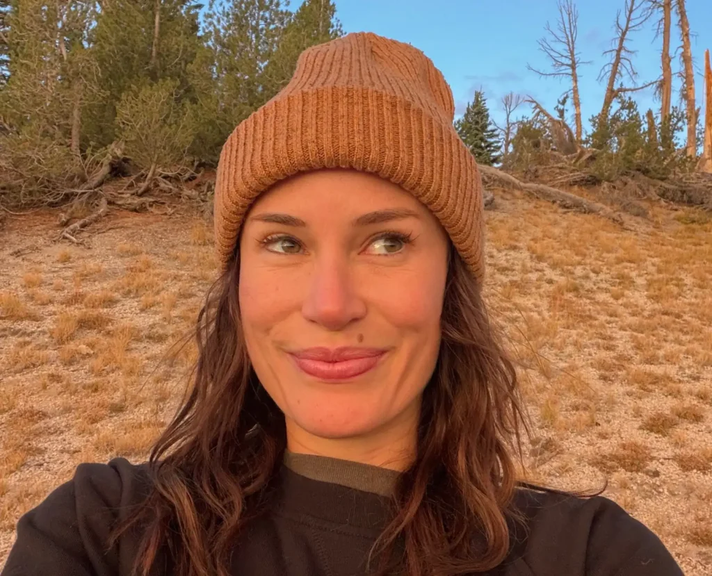 Woman in brown beanie and dark top standing in burnt forest landscape.