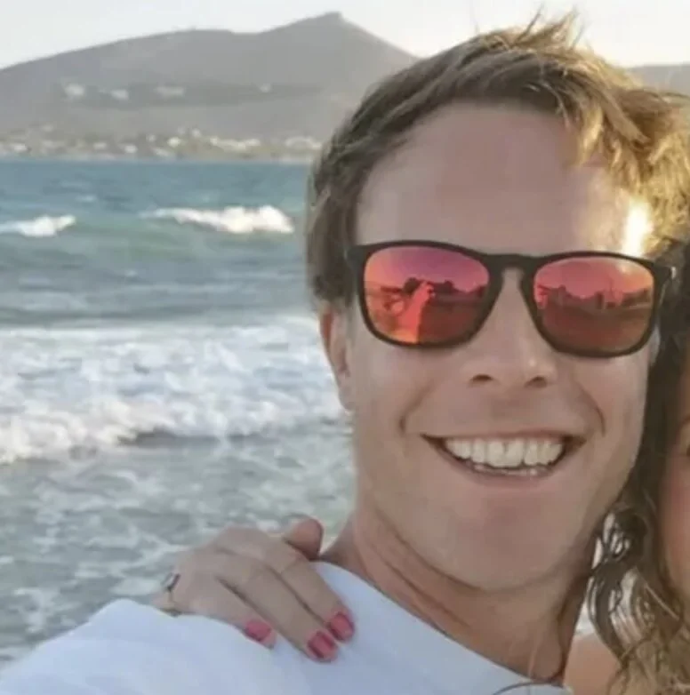 Nathan Vale wearing sunglasses smiling at camera with sea and hillside in background.