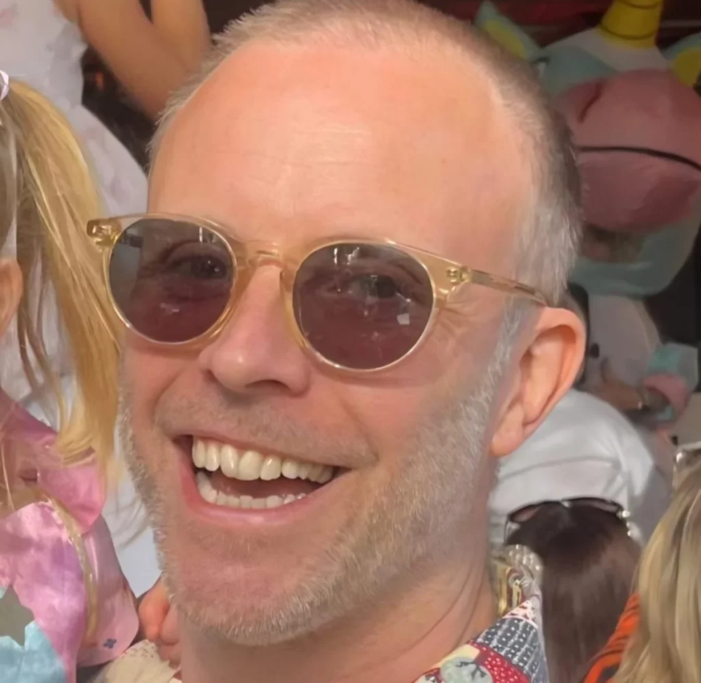 Nathan Vale wearing round glasses and colourful patterned shirt at celebration event.