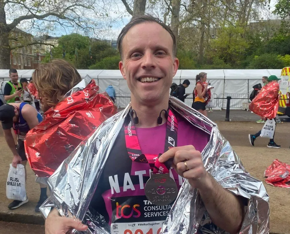 Man wearing silver thermal blanket and race medal number 2011 at outdoor event.