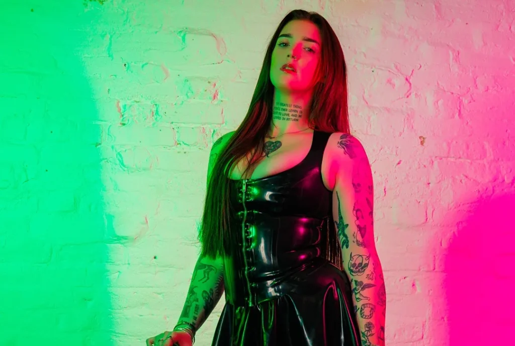 Woman in black latex dress under green and pink neon lighting.